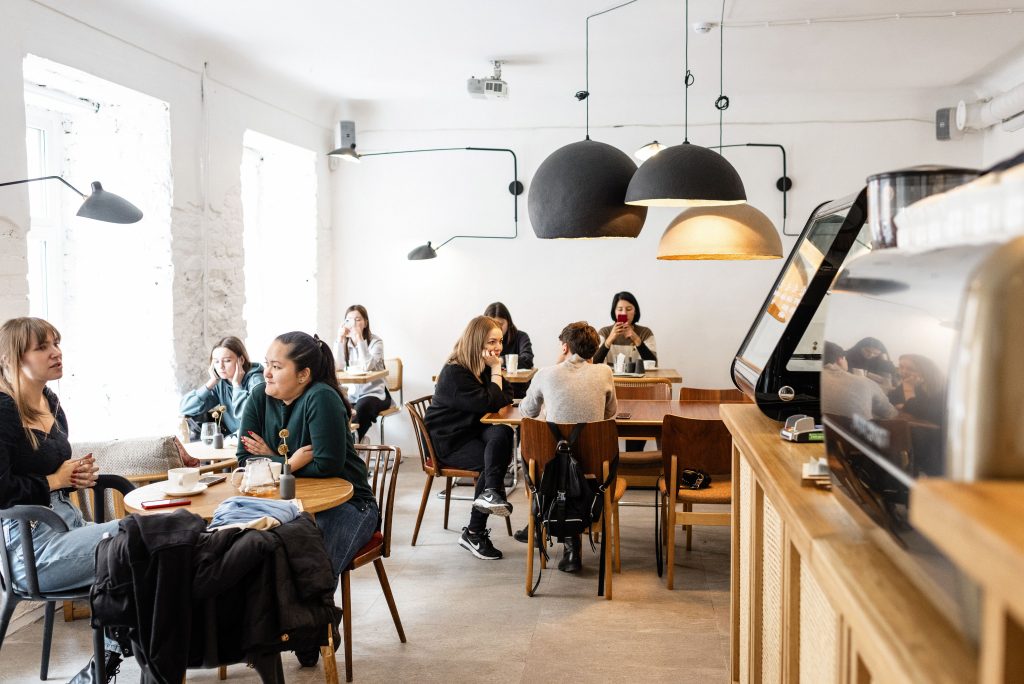 Group of multiracial friends in casual wear sitting at wooden tables in cozy cafeteria near cash box and big lamps while conversing